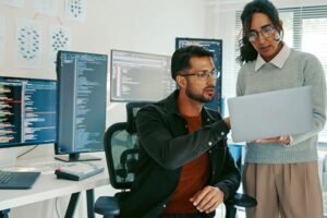 Man seated at a desk pointing to a laptop held by a woman, both reviewing code in a modern workspace with multiple monitors displaying programming interfaces.