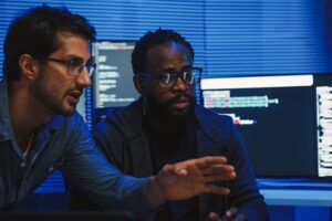 Two men in a dark, tech-lit room collaborating in front of computer monitors displaying code. One gestures toward the screen while the other listens attentively.