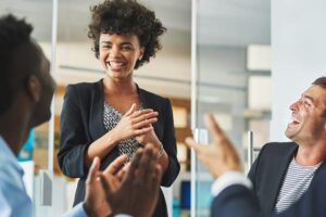 Smiling businesswoman receiving applause from colleagues during a meeting in a modern office.