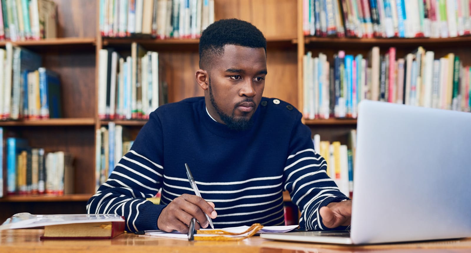 Student in a striped sweater studying at a library table, writing in a notebook while looking at a laptop.