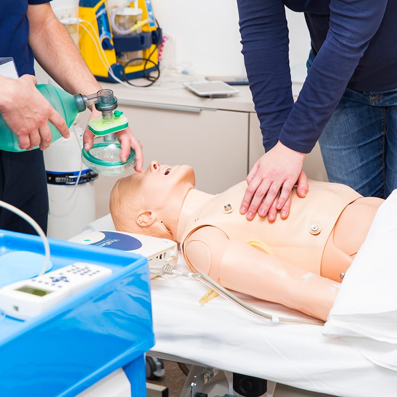 Two individuals perform CPR training on a medical mannequin, with one person administering chest compressions and the other using a manual resuscitator mask for simulated ventilation.