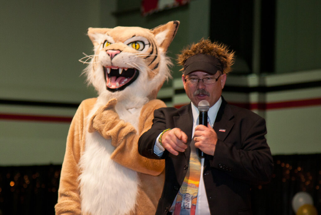 Terry Goerger speaking into a microphone; the Wildcat mascot stands behind him.