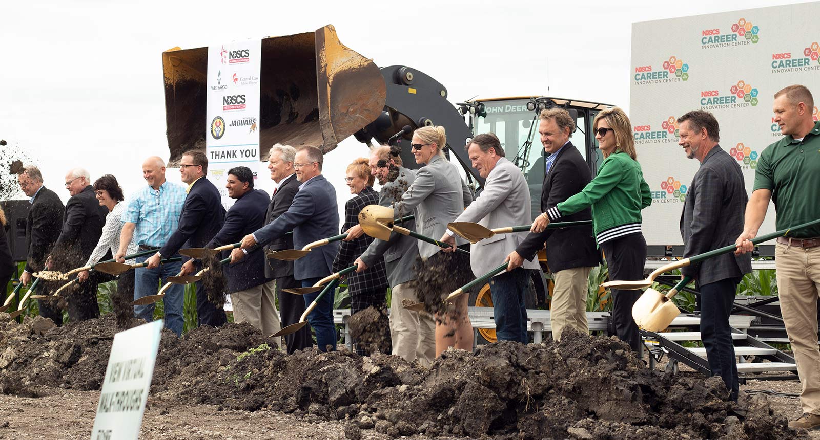 A long line of officials and partners stand in front of a construction site, each holding a gold shovel and lifting soil as part of a ceremonial groundbreaking. Heavy equipment sits behind them along with event signage for the NDSCS Career Innovation Center.