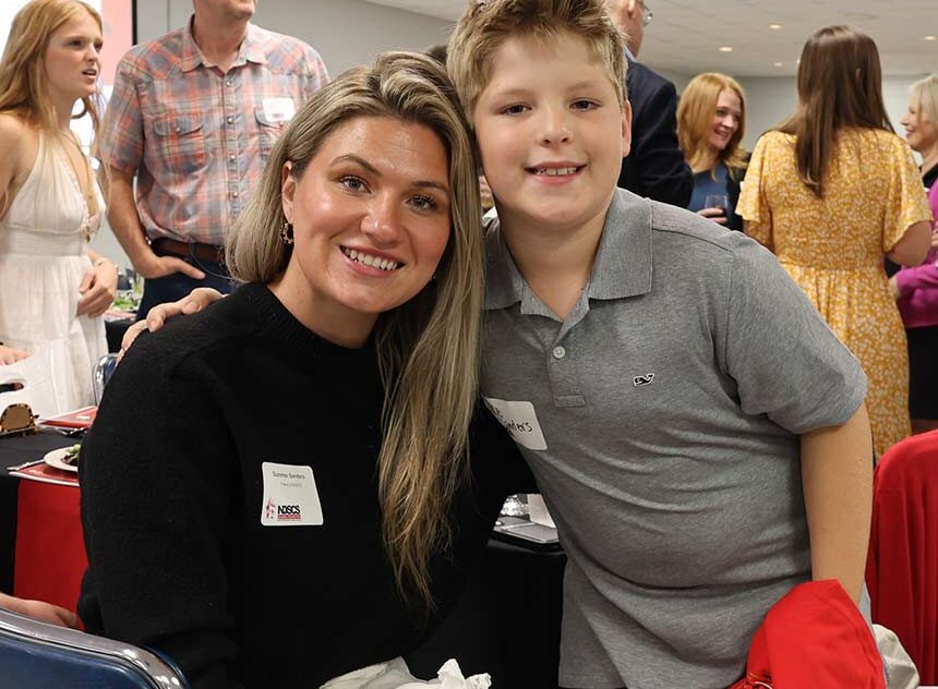 Smiling woman seated at a table with a young boy leaning beside her at a crowded indoor event, with other people talking and mingling in the background.