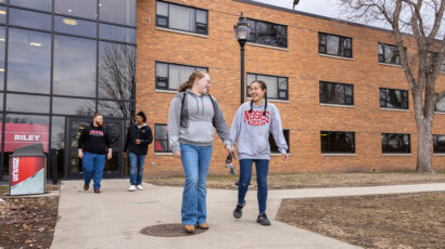 Two female students wearing gray sweatshirts and jeans walking on campus sidewalk. Two male students dressed in black tops and jeans walk behind them.