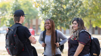 Three students wearing backpacks stand and talk outdoors on campus.