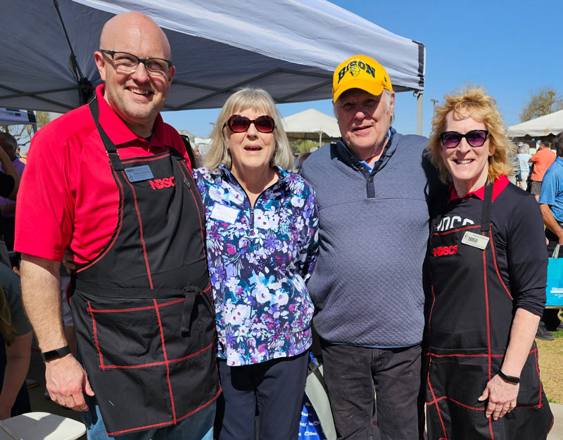 Four adults stand together outdoors under a canopy, smiling at the camera. Two people wear black NDSCS aprons, and the other two stand between them, one wearing a floral top and the other a yellow Bison cap.