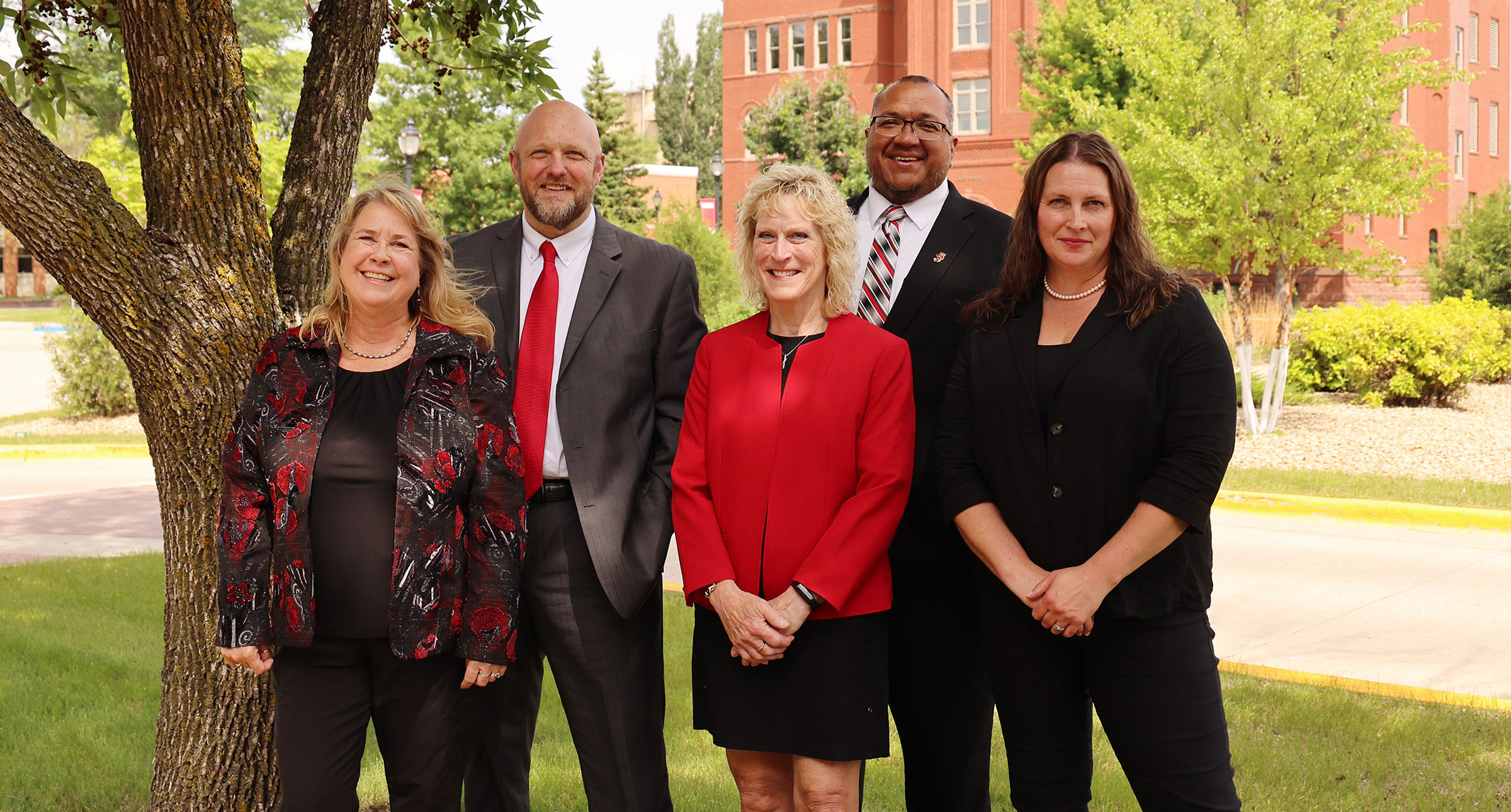 Alumni team members pose outdoors on campus with buildings and trees behind them.