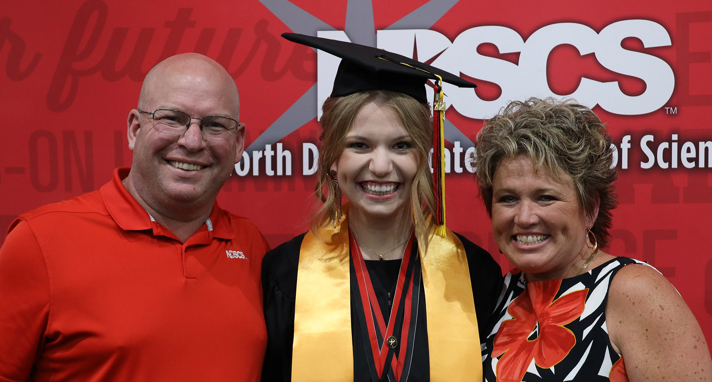 Female graduate wearing a cap & gown with multiple honors poses between a man and woman in front of a red NDSCS backdrop.