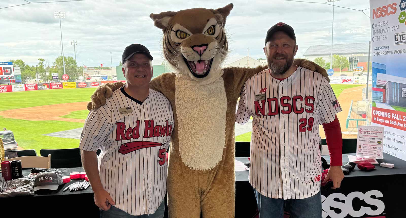 Two men in white pinstripe baseball jerseys stand on either side of a wildcat mascot inside a stadium suite, all smiling with arms around each other. A baseball field is visible behind them along with an NDSCS promotional table and signage.