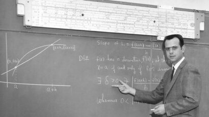 Black and white photo of a math instructor standing at a chalkboard, pointing at a calculus equation about derivatives. A large handwritten graph is on the left side of the board, and a long slide rule hangs above it.