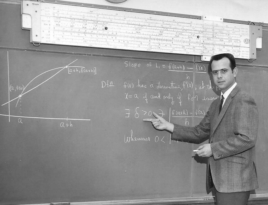 Black and white photo of a math instructor standing at a chalkboard, pointing at a calculus equation about derivatives. A large handwritten graph is on the left side of the board, and a long slide rule hangs above it.