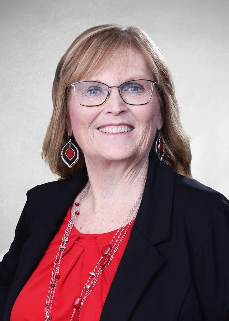 Smiling woman with glasses wearing a black blazer, red top, and matching red jewelry, posed against a light studio backdrop.