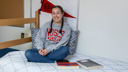Female student wearing a red and gray NDSCS sweatshirt sits on a bed in a dorm room