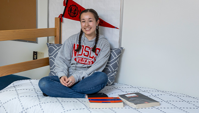 Female student wearing a red and gray NDSCS sweatshirt sits on a bed in a dorm room