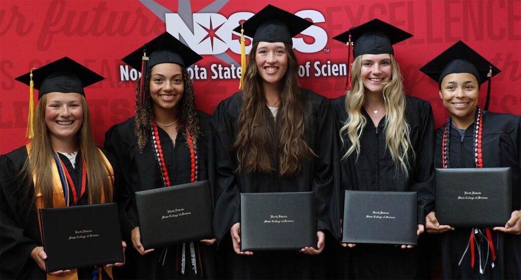 Five graduates in caps and gowns standing in a row, smiling and holding their diploma covers in front of a red backdrop.