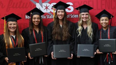 Five graduates in caps and gowns standing in a row, smiling and holding their diploma covers in front of a red backdrop.