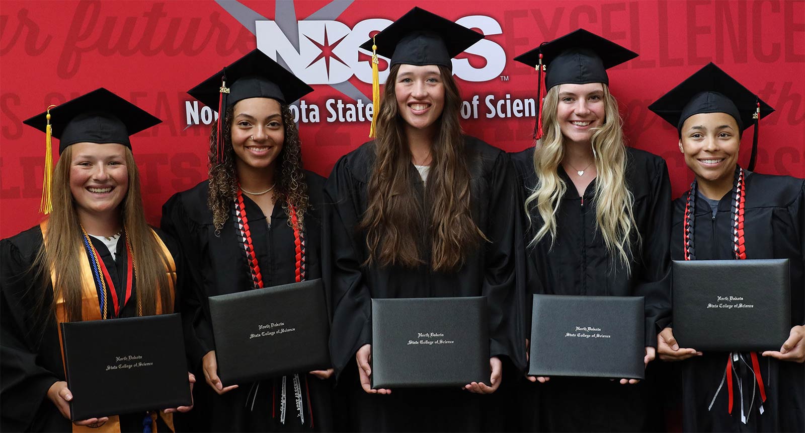 Five graduates in caps and gowns standing in a row, smiling and holding their diploma covers in front of a red backdrop.